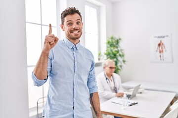 Poster - Hispanic man at doctor clinic smiling with an idea or question pointing finger with happy face, number one