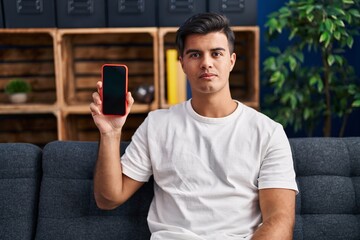 Poster - Hispanic man holding smartphone showing blank screen thinking attitude and sober expression looking self confident