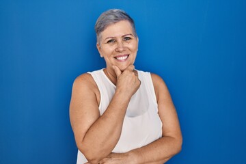 Poster - Middle age caucasian woman standing over blue background looking confident at the camera smiling with crossed arms and hand raised on chin. thinking positive.