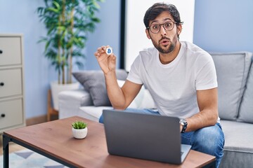 Wall Mural - Handsome latin man holding virtual currency bitcoin using laptop scared and amazed with open mouth for surprise, disbelief face