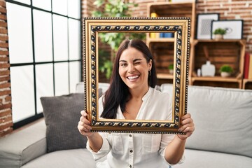 Poster - Young hispanic woman holding empty frame winking looking at the camera with sexy expression, cheerful and happy face.