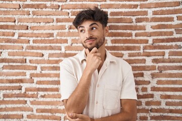 Canvas Print - Arab man with beard standing over bricks wall background looking confident at the camera smiling with crossed arms and hand raised on chin. thinking positive.