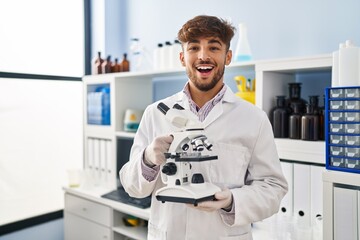 Wall Mural - Arab man with beard working at scientist laboratory holding microscope smiling and laughing hard out loud because funny crazy joke.
