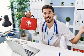Wall Mural - Young hispanic doctor man holding first aid kit celebrating achievement with happy smile and winner expression with raised hand