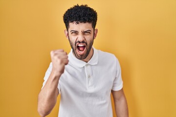 Wall Mural - Arab man standing over yellow background angry and mad raising fist frustrated and furious while shouting with anger. rage and aggressive concept.