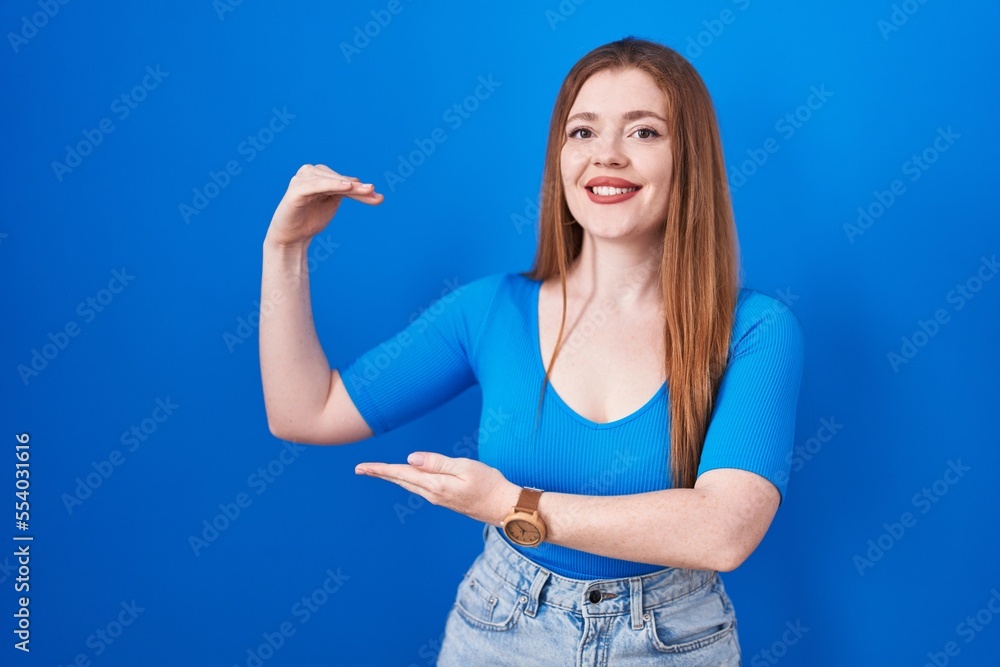 Redhead woman standing over blue background gesturing with hands showing big and large size sign, measure symbol. smiling looking at the camera. measuring concept.