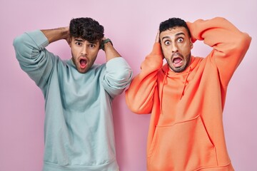 Wall Mural - Young hispanic gay couple standing over pink background crazy and scared with hands on head, afraid and surprised of shock with open mouth
