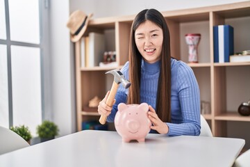 Canvas Print - Chinese young woman holding hammer and piggy bank winking looking at the camera with sexy expression, cheerful and happy face.