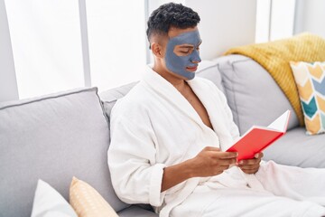 Wall Mural - Young latin man relaxed on sofa with facial mask treatment reading book at home