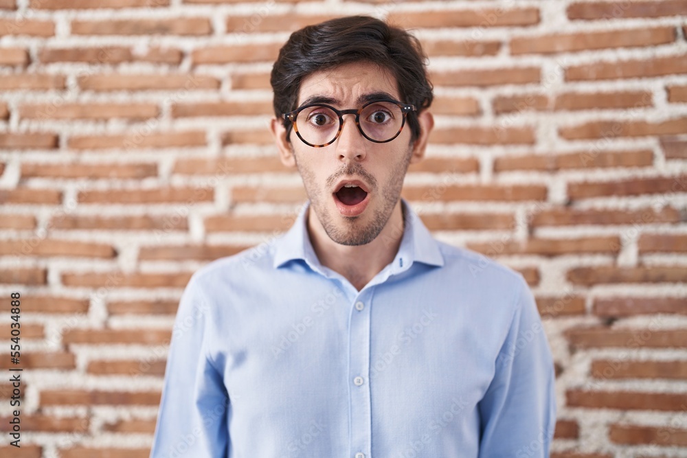 Young hispanic man standing over brick wall background afraid and shocked with surprise and amazed expression, fear and excited face.