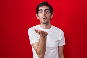 Poster - Young hispanic man standing over red background looking at the camera blowing a kiss with hand on air being lovely and sexy. love expression.