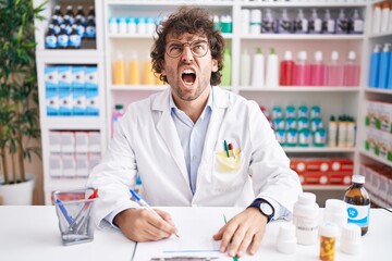 Wall Mural - Hispanic young man working at pharmacy drugstore angry and mad screaming frustrated and furious, shouting with anger. rage and aggressive concept.