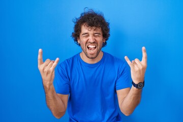 Poster - Hispanic young man standing over blue background shouting with crazy expression doing rock symbol with hands up. music star. heavy music concept.