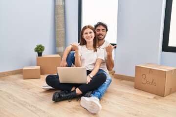 Canvas Print - Young couple using laptop at new home pointing to the back behind with hand and thumbs up, smiling confident