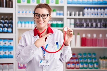 Poster - Young hispanic woman with red hair working at pharmacy drugstore using stethoscope smiling happy pointing with hand and finger