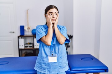 Poster - Young hispanic woman wearing physiotherapist uniform standing at clinic tired hands covering face, depression and sadness, upset and irritated for problem