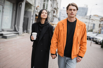 Stylish man holding hand of asian girlfriend with coffee to go on urban street.