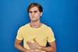 © Krakenimages.com - Young man standing over blue background with hand on stomach because nausea, painful disease feeling unwell. ache concept.