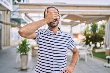 Canvas Print - Young hispanic man with beard outdoors at the city smiling and laughing with hand on face covering eyes for surprise. blind concept.