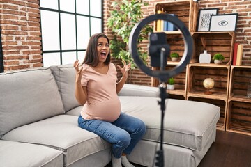 Wall Mural - Young pregnant woman recording vlog tutorial with smartphone at home crazy and mad shouting and yelling with aggressive expression and arms raised. frustration concept.