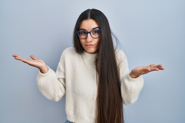 Canvas Print - Young hispanic woman wearing casual sweater over blue background clueless and confused expression with arms and hands raised. doubt concept.