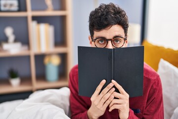 Sticker - Young caucasian man covering face with book sitting on bed at bedroom