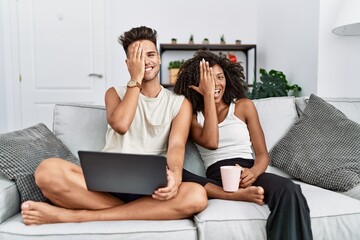 Sticker - Young interracial couple using laptop at home sitting on the sofa covering one eye with hand, confident smile on face and surprise emotion.