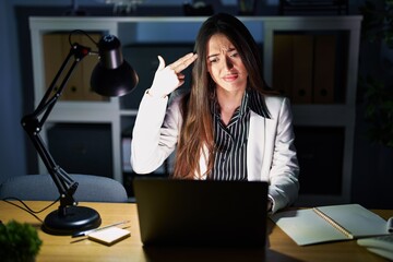 Sticker - Young brunette woman working at the office at night with laptop shooting and killing oneself pointing hand and fingers to head like gun, suicide gesture.