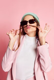 Fototapeta Do pokoju - Portrait of young beautiful girl in a suit, hat and sunglasses posing over pink background. Good mood