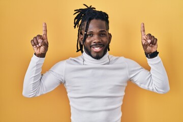 Poster - African man with dreadlocks wearing turtleneck sweater over yellow background smiling amazed and surprised and pointing up with fingers and raised arms.