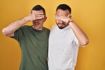 Wall Mural - Homosexual couple standing over yellow background covering eyes with hand, looking serious and sad. sightless, hiding and rejection concept