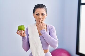 Canvas Print - Young woman wearing sportswear and towel eating green apple covering mouth with hand, shocked and afraid for mistake. surprised expression