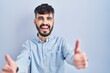 Young hispanic man with beard standing over blue background approving doing positive gesture with hand, thumbs up smiling and happy for success. winner gesture.