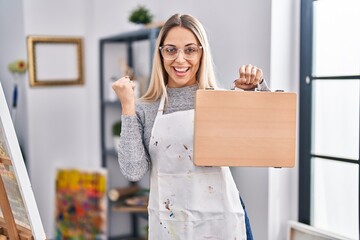 Poster - Young blonde painter woman holding wooden case at art studio screaming proud, celebrating victory and success very excited with raised arms