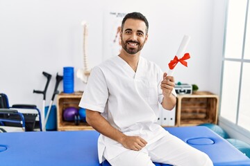 Canvas Print - Young hispanic man wearing physiotherapist uniform holding diploma at clinic