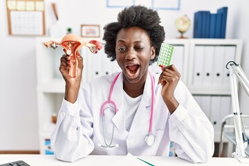 Canvas Print - African doctor woman holding anatomical female genital organ and birth control pills winking looking at the camera with sexy expression, cheerful and happy face.