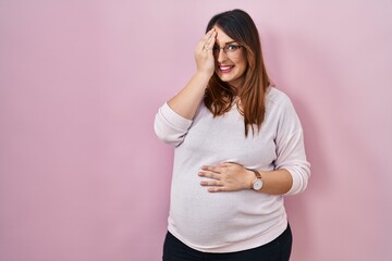 Poster - Pregnant woman standing over pink background covering one eye with hand, confident smile on face and surprise emotion.