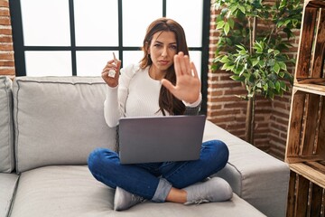 Poster - Young brunette woman using laptop holding keys of new home with open hand doing stop sign with serious and confident expression, defense gesture