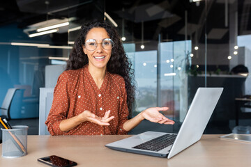 Wall Mural - Portrait of happy and successful businesswoman, Hispanic woman smiling and looking at camera sitting at desk inside modern office building, using laptop at work.