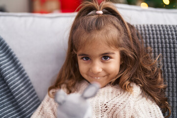 Canvas Print - Adorable hispanic girl holding doll sitting on sofa by christmas tree at home