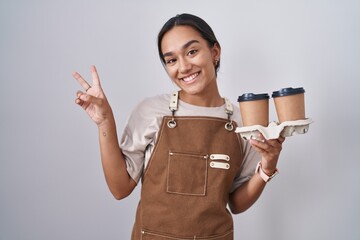 Wall Mural - Young hispanic woman wearing professional waitress apron holding coffee smiling looking to the camera showing fingers doing victory sign. number two.
