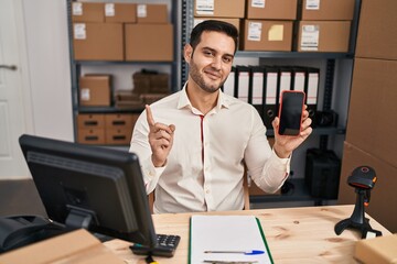 Canvas Print - Young hispanic man with beard working at small business ecommerce showing smartphone screen smiling happy pointing with hand and finger to the side