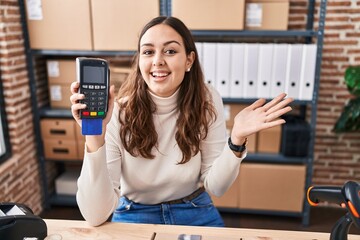 Sticker - Young hispanic woman working at small business ecommerce holding dataphone celebrating victory with happy smile and winner expression with raised hands