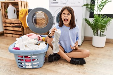 Poster - Young hispanic girl doing laundry holding socks angry and mad screaming frustrated and furious, shouting with anger. rage and aggressive concept.