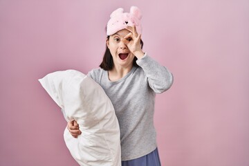 Poster - Woman with down syndrome wearing sleeping mask hugging pillow doing ok gesture shocked with surprised face, eye looking through fingers. unbelieving expression.