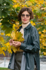 Wall Mural - A woman in leather clothes stands against the backdrop of an autumn maple square and holds maple leaves in her hands