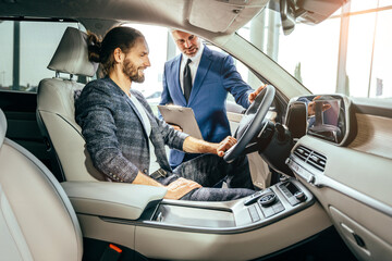 Cheerful handsome man sitting in a new car at the dealership smiling talking to a mature salesman while choosing a new automobile buying buyer selling seller communication experience advice service.
