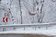 Fragment winter road with mirror installed near sharp turn fenced with iron bump stop with red arrow indicating direction. Northern track with gray sleet on way and snowbound forest behind roadside 