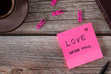 Wall Mural - Love never fails, handwritten biblical verse on pink note with cup of coffee and holy bible book on wooden background. Top table view.