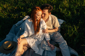 Wall Mural - smiling ginger young woman in dress and romantic man in love sitting hugging together on beautiful g